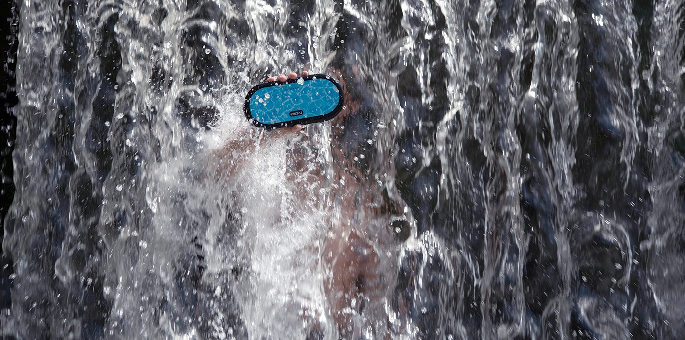 Man holding Zeppy blue wireless Bluetooth speaker under a waterfall.